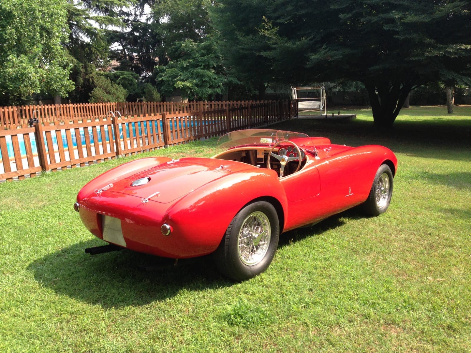 Classic red open-top sports car with wire wheels parked on grass near a pool.